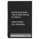 Small black spiral notebook laser etched with 'We're Building The Plane While Flying It. And Other B.S. Phrases' design